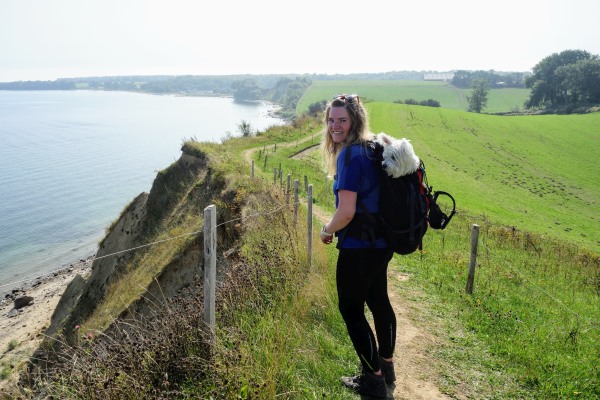 Vrouw wandelt langs de Deense kust, met een hond in haar rugzak.
